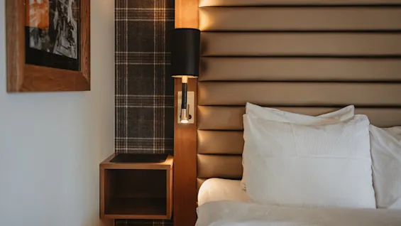 Et indbydende hotelværelse med en plys seng med polstret sengegavl, blødt hvidt sengetøj og en varm sengelampe til læsning.