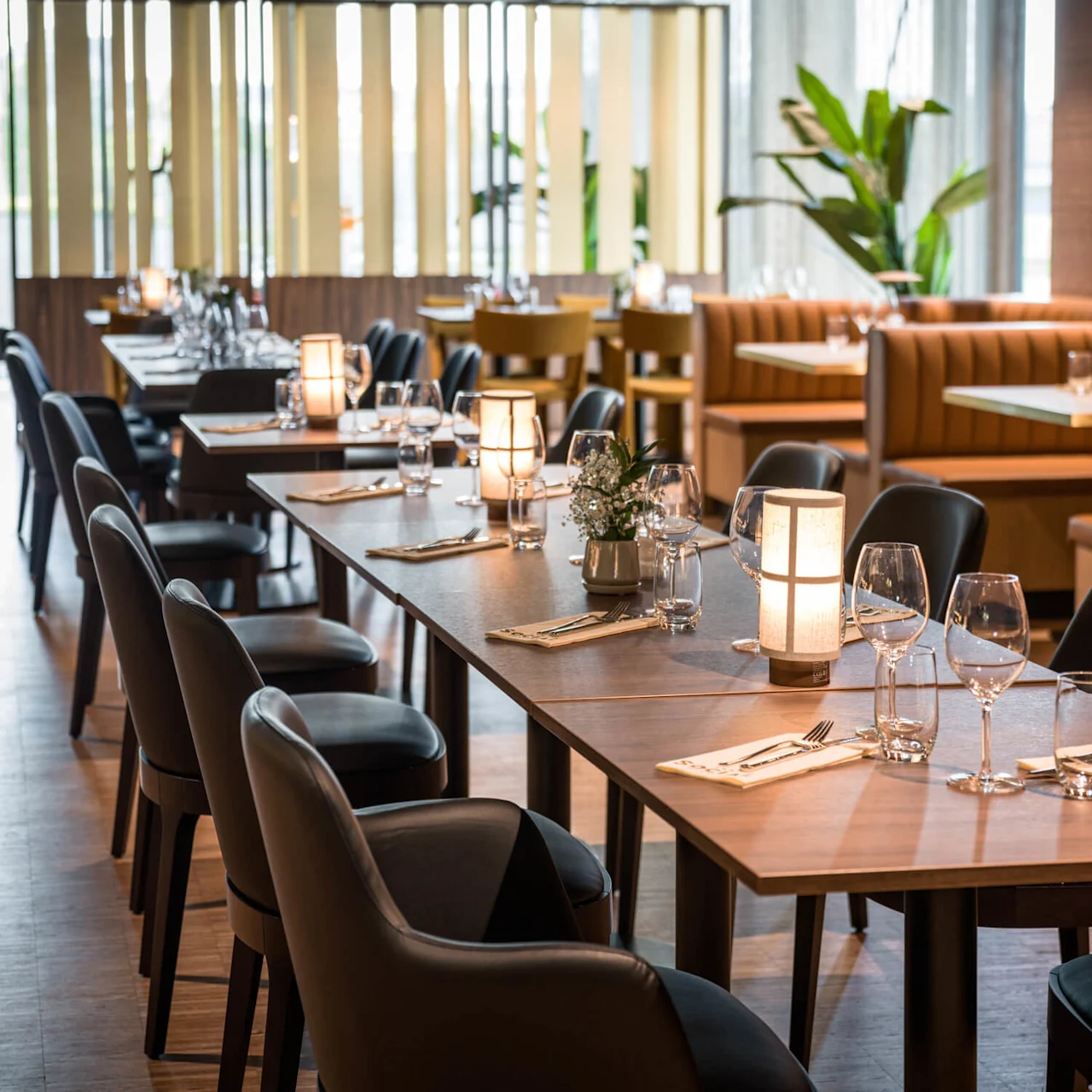 Et trebord med vinglass, lys og servietter i en elegant restaurant, omgitt av skinnstoler og båsseter, med planter i bakgrunnen.