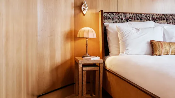 Et koselig soveromshjørne med seng, nattbord og lampe i et rom med trepaneler og fiskebensmønstret gulv samt et mønstret teppe.