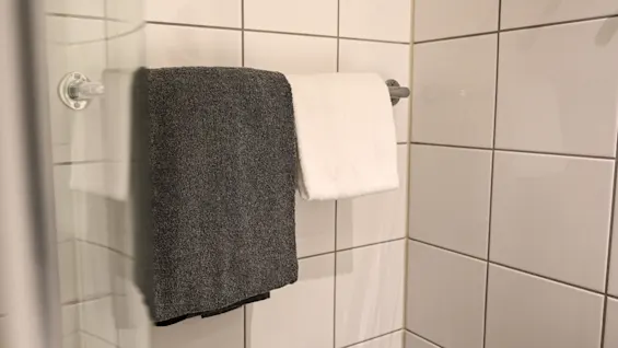 Två handdukar, en grå och en vit, hänger prydligt på en metallstång mot en vit kaklad badrumsvägg.