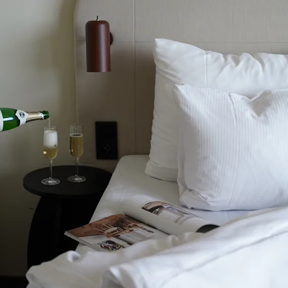 Champagne hälls upp i glas bredvid en bäddad hotellsäng med vita lakan och en tidning.
