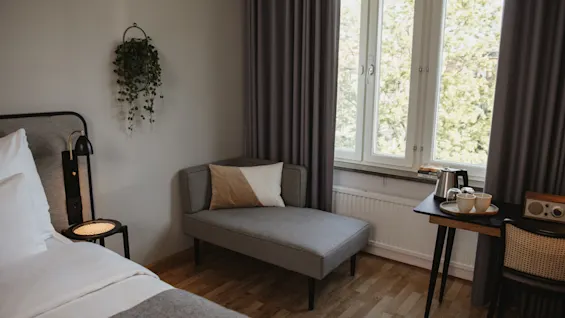 Seng med hvitt sengetøy ved siden av en grå chaiselong i et rom med tregulv. Bord med kaffesett nær vinduet med lyse gardiner, plante dekorerer veggen.