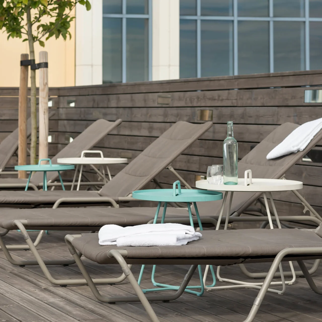 Sun loungers are arranged neatly on a wooden deck by a wooden fence, with small tables holding bottles and glasses, providing a relaxing outdoor setting with natural colors.