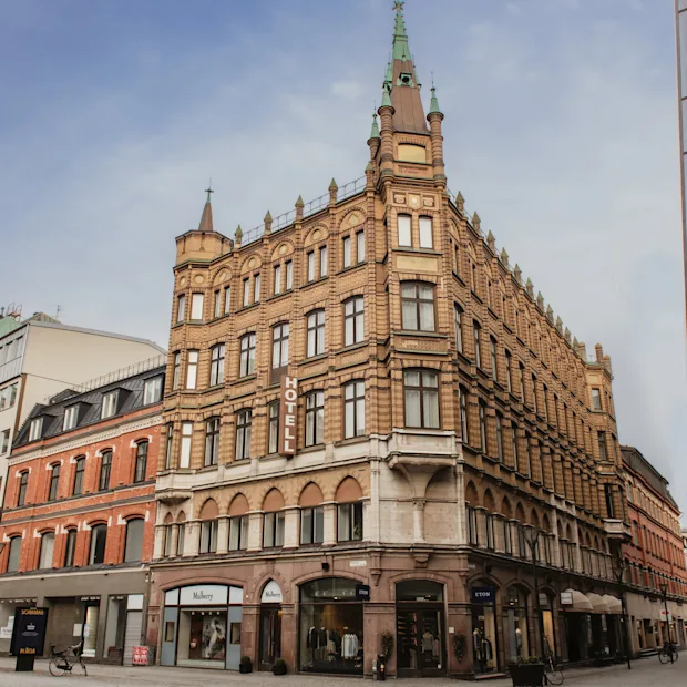 Home Hotel Baltzar. A grand, ornate brick building with a spire stands on a city street, with other buildings nearby. Text: HOTELL.