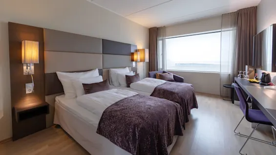 Two neatly made single beds with brown blankets are side-by-side in a modern hotel room featuring a large window, chairs, and a desk with a kettle and cups.
