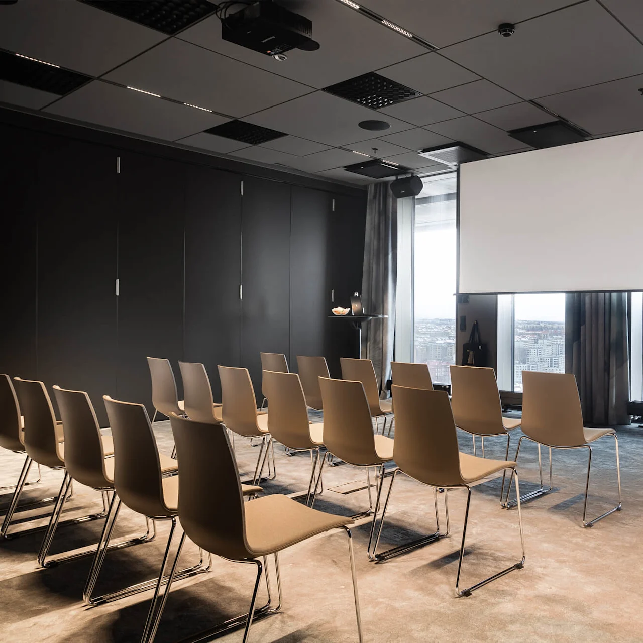 Rader av tomma stolar vända mot en stor projektorduk i ett konferensrum med fönster från golv till tak som visar en stadsbild. Ett litet sidobord har en bärbar dator och vattenflaska.