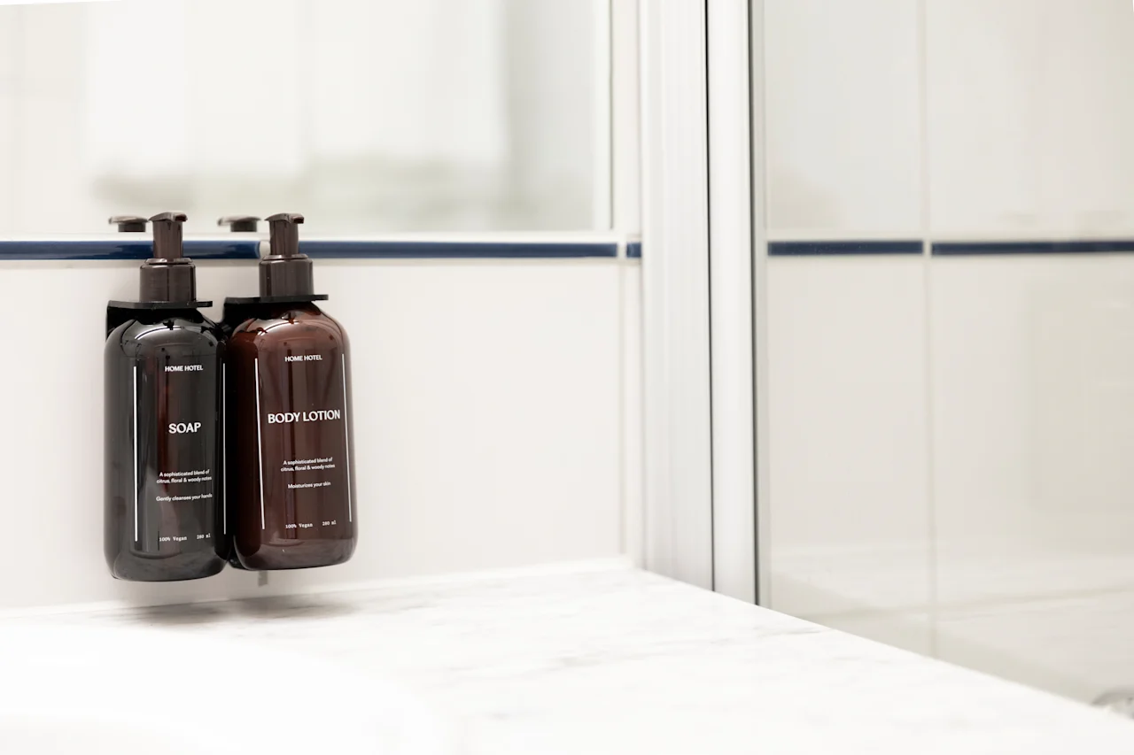 Kaksi annostelijapulloa, toinen SAIPPUA ja toinen BODY LOTION, valkoisella kaakeloidulla kylpyhuoneen seinällä.