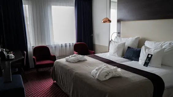 En prydligt bäddad hotellsäng med morgonrockar har ett täcke och kuddar i ett rum med stoppade stolar och mjukt ljus från en hängande lampa.
