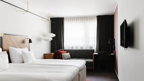 Ett snyggt hotellrum med två enkelsängar, en soffa, ett skrivbord och en väggmonterad TV, vilket antyder komfort och funktionalitet.