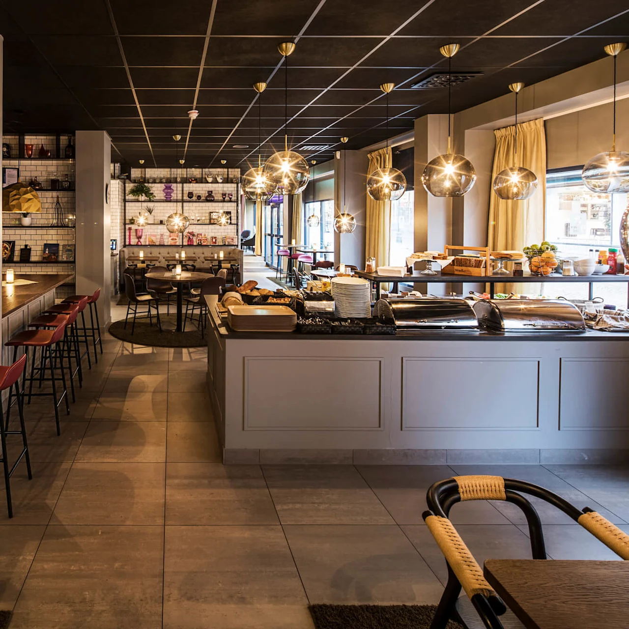A cafe interior with tables, chairs, and modern pendant lighting; a buffet area with bread and fruits, providing a cozy atmosphere.