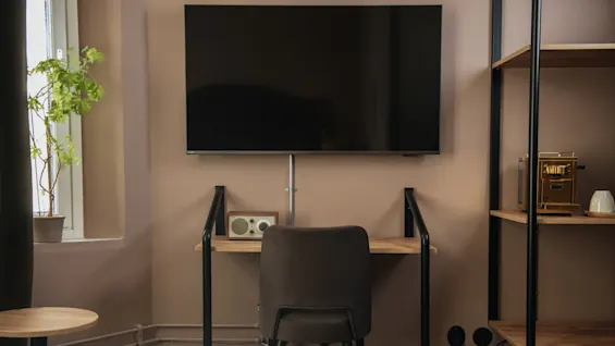 A wall-mounted TV is above a small desk with a chair, radio, and potted plant by a window. Shelves hold a coffee machine and cup nearby.