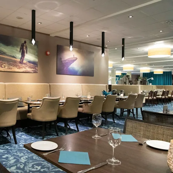 Restaurant dining area with beige chairs arranged around tables set with glassware and napkins. Large adventure-themed images hang on the wall, and soft lighting illuminates the spacious interior.