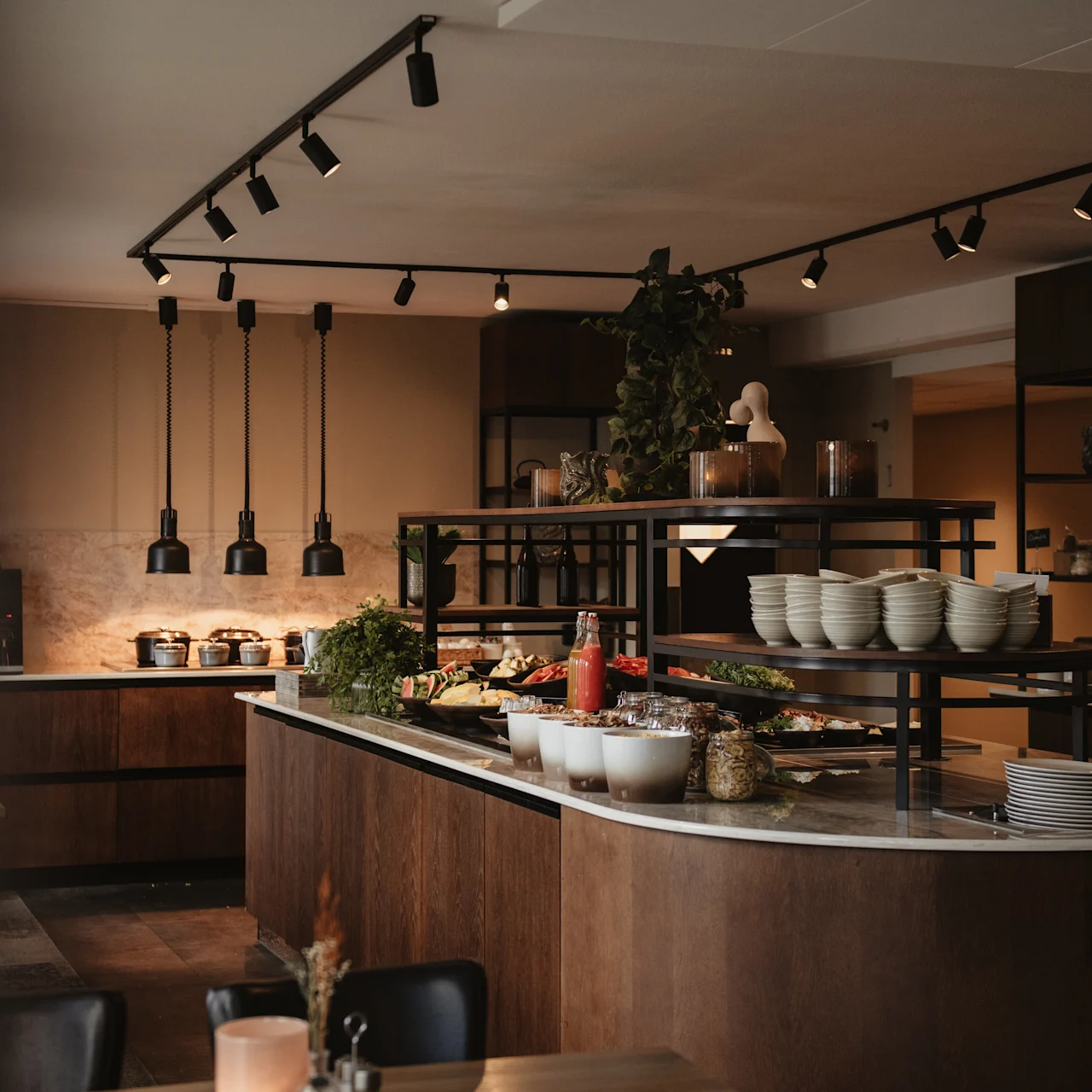 A modern buffet table displays various dishes and bowls under pendant lights, in a contemporary dining area with wooden and metal accents.