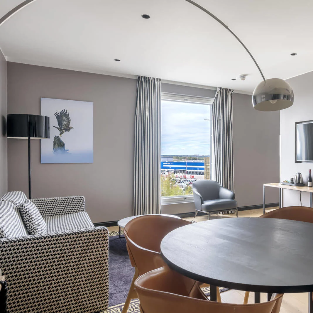 Circular table surrounded by leather chairs stands in a modern hotel room. A striped sofa, large TV, and bedside area are present. Window reveals an exterior view.