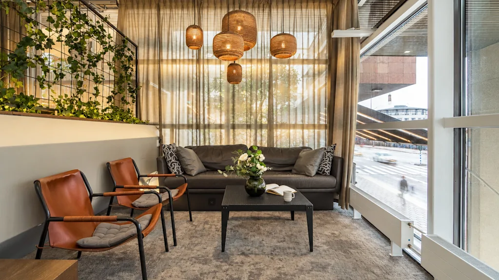 Cozy hotel lobby lounge with a dark sofa, brown leather armchairs, a coffee table, and hanging wicker lamps by a window.
