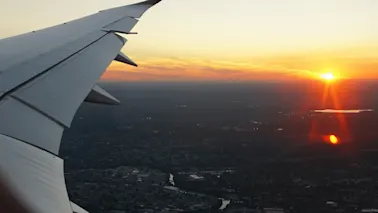 Flyvinge strekker seg ut, svevende over et spredt bylandskap. En levende solnedgang farger himmelen med oransje nyanser, med refleksjoner på et fjernt vann.