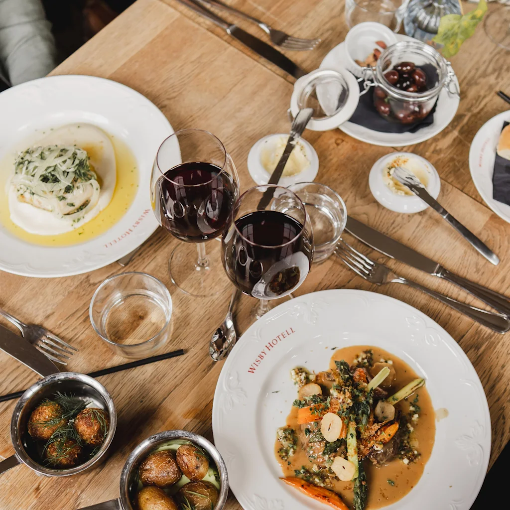 Swedish: Två tallrikar med gourmetmat står på ett träbord, ackompanjerade av röda vinglas och små skålar, i en elegant restaurangmiljö.