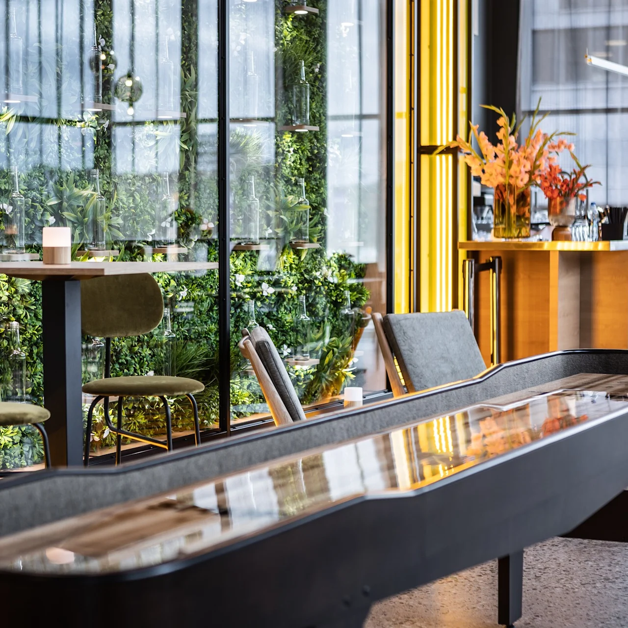 Ett shuffleboard-bord sträcker sig över en modern, stilren interiör. Omgivande är gröna växter bakom glas, höga pallar och en bar med livfulla blomsterarrangemang, vilket skapar en samtida atmosfär.