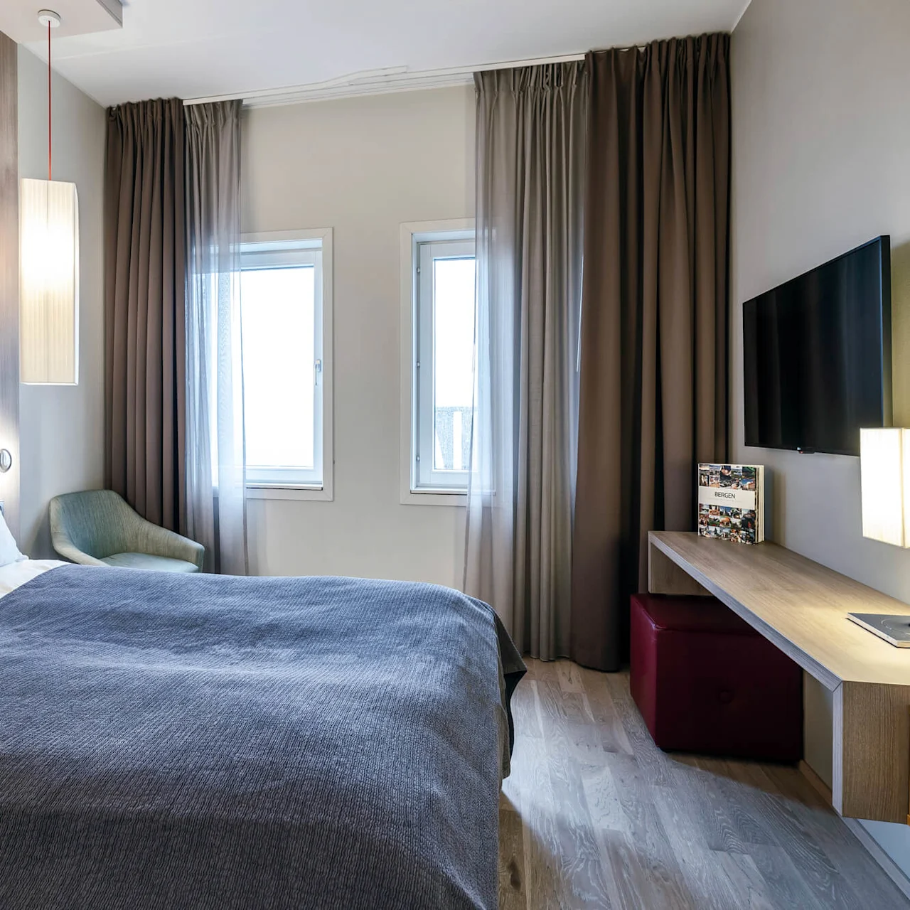 Et koselig hotellrom har en pent oppredd seng med et grått teppe ved siden av et lite bord med stol, en veggmontert TV og store vinduer med gardiner.
