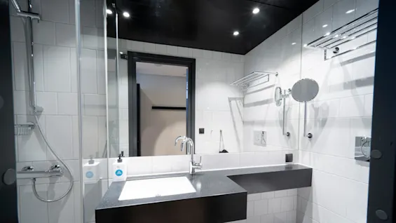 A modern bathroom with a sleek sink and faucet; mirrors and white tiled walls surround. A glass-enclosed shower and towel racks are visible.