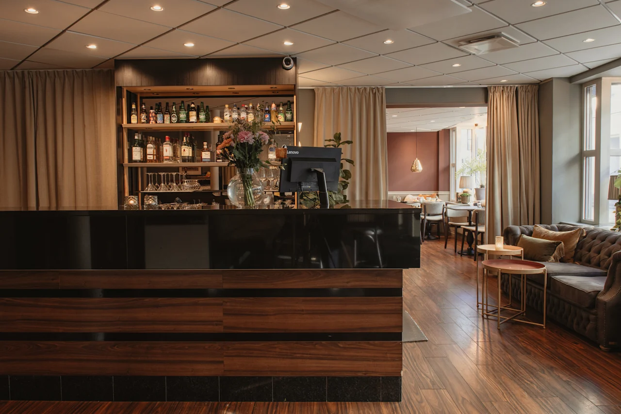 Lämmin ja kutsuva hotellibaari ja lounge-alue, jossa on tumma puinen baari, pulloilla täytetyt hyllyt ja mukava sohva.