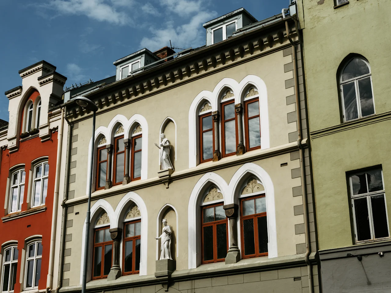Udsmykkede bygningsfacader med buede vinduer og dekorative statuer under en blå himmel. En blanding af arkitektoniske stilarter og farver.