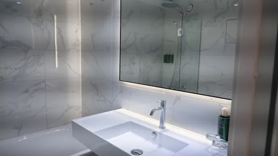 Et moderne badeværelse med hvid vask, kromhane og stort spejl, der reflekterer en bruser. Marmormønstrede fliser.