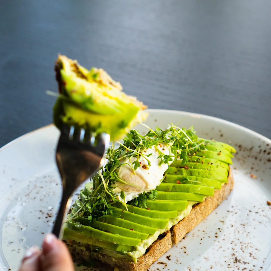 Avokadotoast, toppet med skivet avokado, krem og spirer, serveres på en flekkete tallerken. En hånd holder en gaffel med en bit av toasten i forgrunnen.