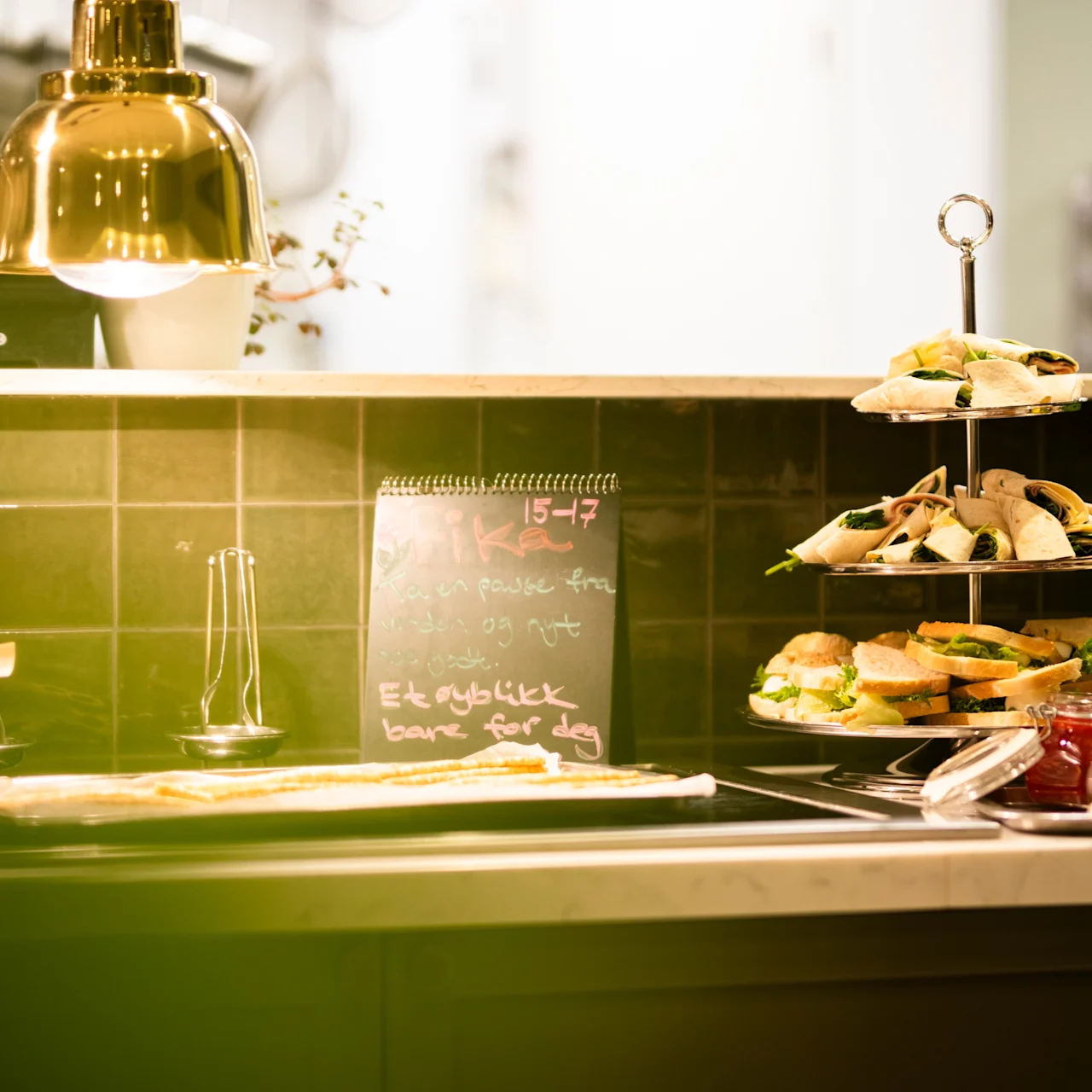 A tiered stand holds sandwiches on a kitchen counter lined with various jars. A chalkboard is in the background with text: "15-17 Tarte fris Entre Ek øyeblikk bare for deg."