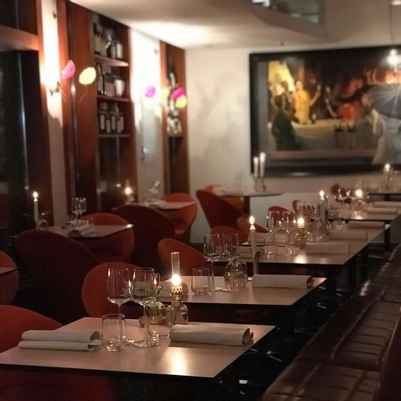 Tables set with glassware and napkins, surrounded by red chairs in a dimly lit restaurant. Wall shelves hold bottles; a large painting and candles add to the ambiance.