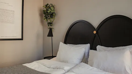 En snyggt bäddad säng med vita kuddar och ett mörkt sänggavel står bredvid en liten, grön krukväxt på ett högt ställ. En inramad konstverk hänger på en beige vägg.