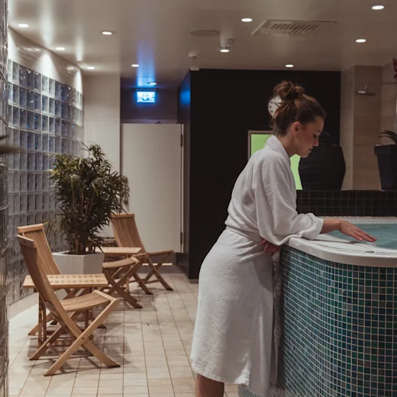 En person i vit badrock testar vattnet i en bubbelpool i ett svagt upplyst spaområde, omgiven av växter och trästolar.