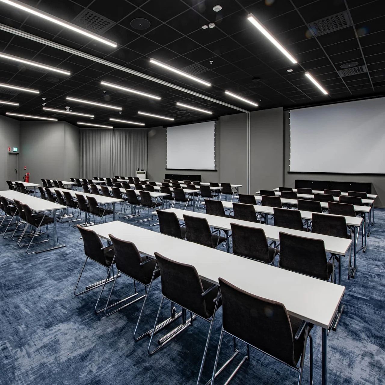 Rows of black chairs and white tables face two large projection screens in a spacious conference room with blue carpet and overhead fluorescent lighting.