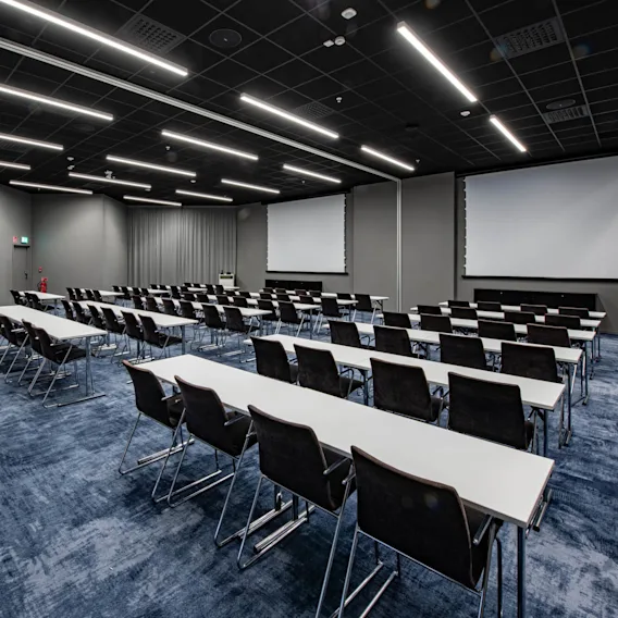 Rader med svarta stolar och vita bord vetter mot två stora projektionsskärmar i ett rymligt konferensrum med blå matta och lysrörsbelysning ovanför.