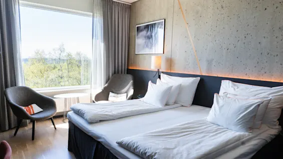 En prydligt bäddad dubbelsäng mot en texturerad vägg, flankeras av två fåtöljer bredvid ett stort fönster med utsikt över träd. Mjuk belysning och en inramad bild kompletterar det moderna rummet.