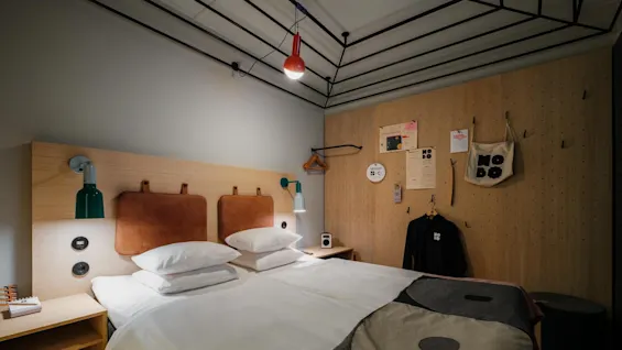 A bed with white linens and brown headboards is in a stylish room. Two lamps and wall decor, including hangers and a bag labeled "Nogo," are attached to wooden walls.