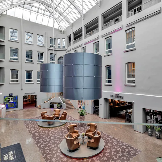 Large cylindrical lamps hang in a spacious atrium with a glass ceiling, surrounded by multiple floors with windows and a bar area labeled "BAR & A LA CARTE."