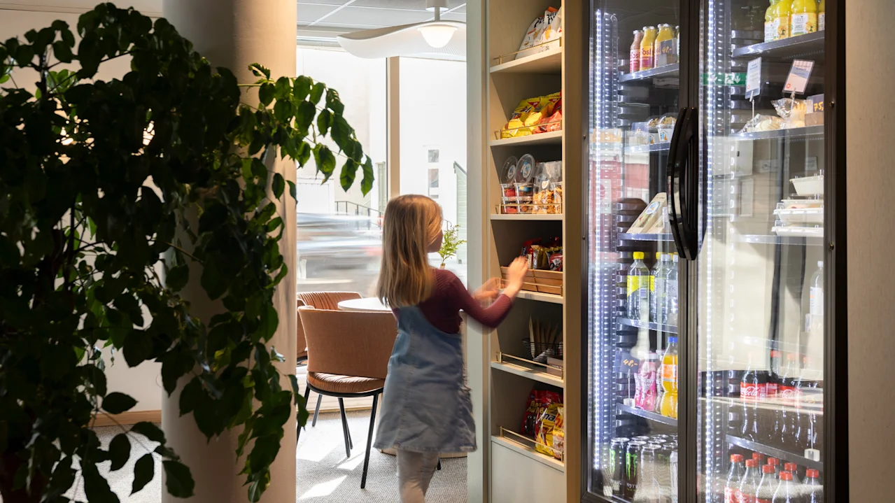 Ett barn sträcker sig efter föremål på snacks-hyllor bredvid ett kylskåp fyllt med drycker. Miljön är ett väl upplyst rum med en stor växt och stolar i bakgrunden.