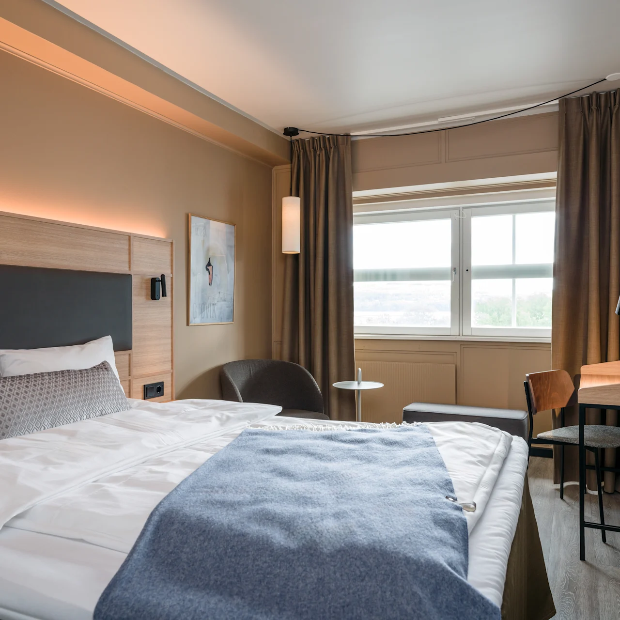 A bed is neatly made with white and blue bedding, situated in a modern hotel room. The room features a desk with a chair, a window with curtains, and wall art.