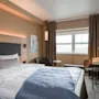 A bed is neatly made with white and blue bedding, situated in a modern hotel room. The room features a desk with a chair, a window with curtains, and wall art.