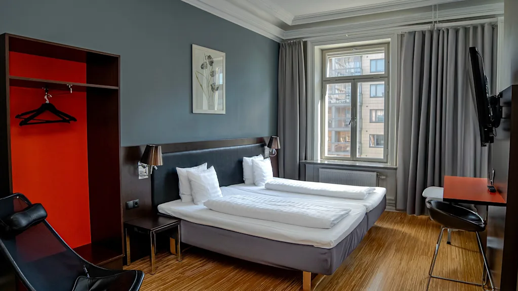Ett modernt hotellrum med två sängar, ett fönster med grå gardiner, en garderob och en liten skrivbordsplats.