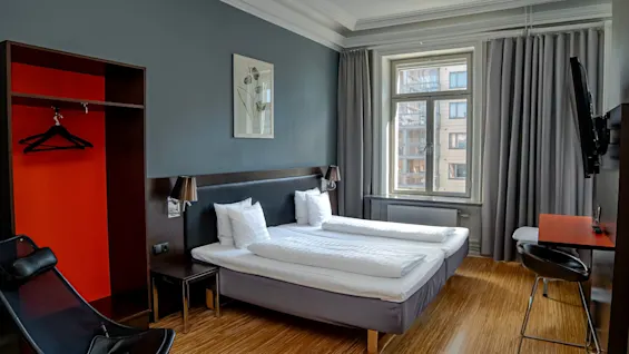 Ett modernt hotellrum med två sängar, ett fönster med grå gardiner, en garderob och en liten skrivbordsplats.