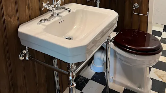 En hvid porcelænsvask med kromarmaturer er monteret på en træpanelvæg ved siden af et toilet. Et spejl hænger over, og gulvet er sort-hvidt ternet.