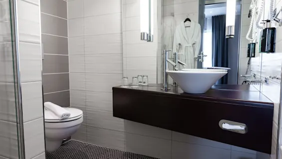 Modern bathroom with a wall-mounted sink and mirror, toilet with folded towel, and glass shelves holding toiletries. Context includes white and gray tiles and a hanging bathrobe on the door.