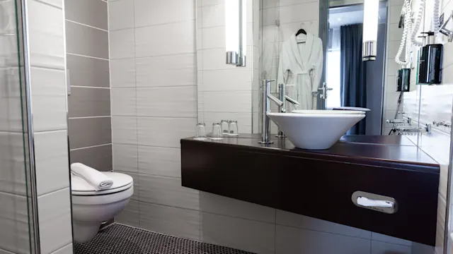 Modern bathroom with a wall-mounted sink and mirror, toilet with folded towel, and glass shelves holding toiletries. Context includes white and gray tiles and a hanging bathrobe on the door.