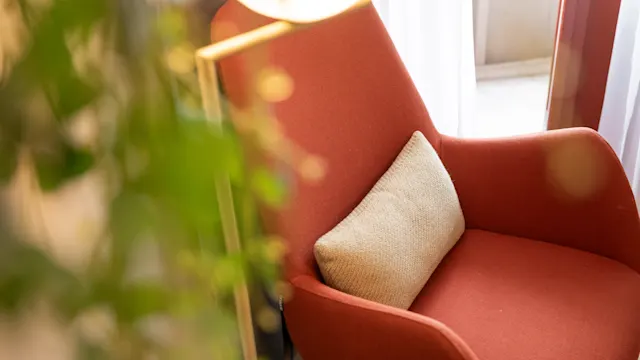 En oransje lenestol holder en beige pute, plassert nær et lyst vindu. Grønt løvverk er delvis synlig i forgrunnen.