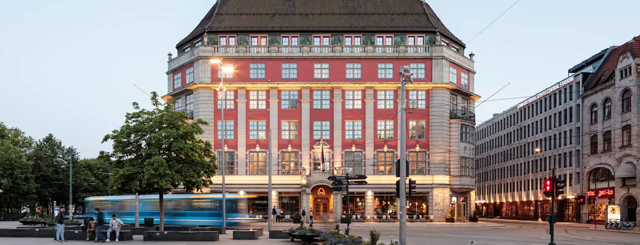 Ett stort, utsmyckat byggnad står på ett torg i skymningen, med en spårvagn som passerar. Text lyder: "CLARION HOTEL THE HUB OSLO CENTRAL STATION." Människor sitter i närheten.