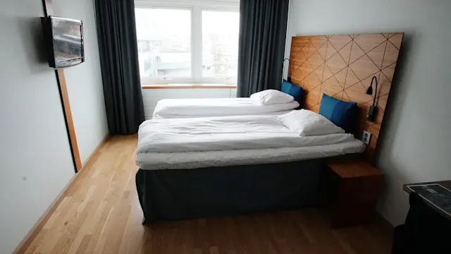 Huolellisesti pedattu valkoinen sänky on asetettu puupaneelin eteen minimalistisessa huoneessa. Taulutelevisio roikkuu seinällä suurten ikkunoiden vieressä, joissa on tummat verhot.