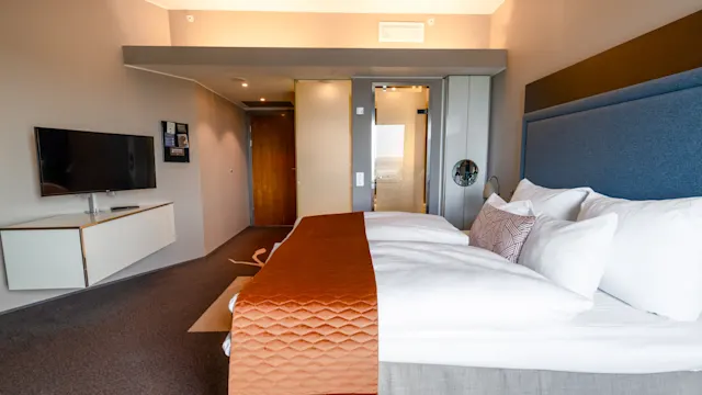 Vuode, jossa on valkoiset lakanat ja oranssi peite, on modernissa hotellihuoneessa. Seinälle kiinnitetty TV ja minimalistiset huonekalut näkyvät, samoin kuin kylpyhuoneen sisäänkäynti.