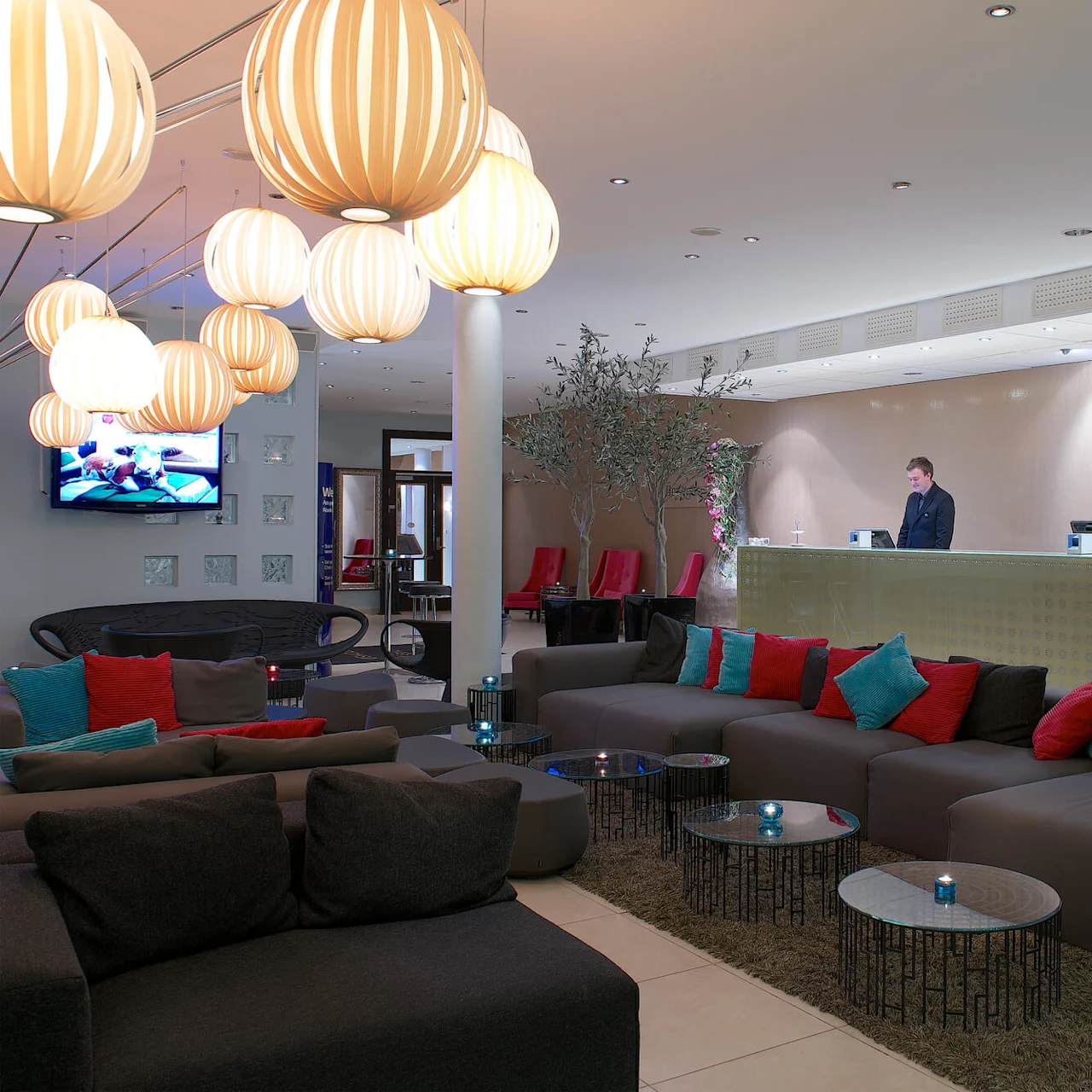 A modern hotel lobby with a reception desk, sofas, decorative cushions, stylish lighting, and a TV on the wall.